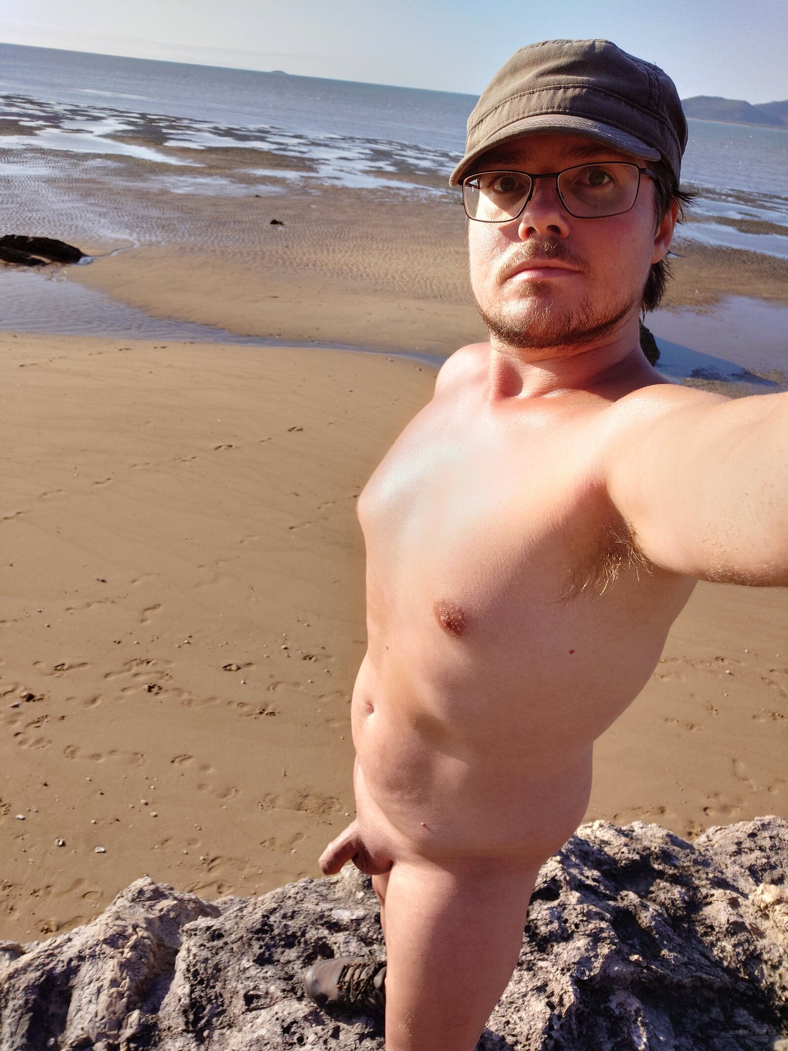 Nude man in cap and glasses selfies on beach rocks with tidal sand flats and sea behind him.