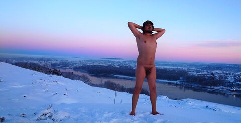 A nude bearded man in a cap and glasses stands barefoot on a snowy hillside above a river town at dusk.